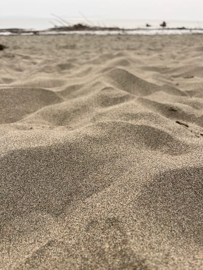 The image shows a close-up, low-angle view of sand dunes on a beach. The sand is light brown and textured, with subtle variations in tone and shadow creating undulating patterns. In the background, a hazy horizon separates the beach from a pale sky, with faint indications of water and some dark, indistinct objects, possibly vegetation or debris, along the shoreline. There are no people or discernible structures in the immediate foreground or midground. The overall impression is one of natural, unoccupied beach landscape. The location is identified as Krasnoturansk, Russia.
