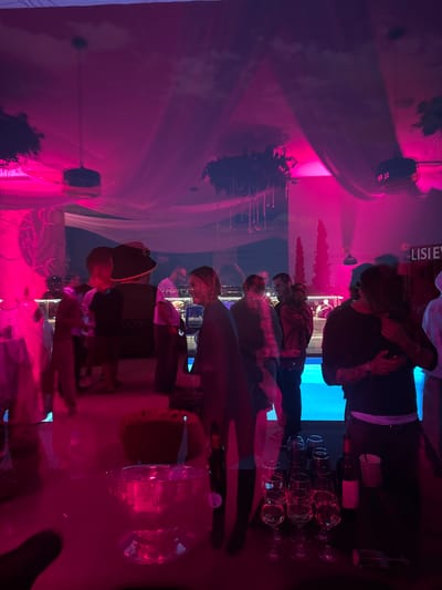 In the city of Mukhatskaro, Georgia, an indoor event is taking place, illuminated by vibrant pink and blue lighting, casting a surreal glow on the scene. The atmosphere appears to be that of a party or social gathering. A group of people are casually mingling, their figures silhouetted against the colored lights. Some are seen in conversation, while others are standing or walking. In the foreground, a table is set with various items. Several wine glasses are arranged, some containing a reddish liquid, suggesting the presence of wine. A bottle of wine, likely red, stands upright next to a red paper cup. An ice bucket filled with ice and possibly beverages sits in the middle of the table. The background reveals a spacious area with draped fabric ceiling decorations, possibly resembling clouds, and plants, creating a festive ambiance. The overall scene suggests a lively social event with a focus on drinks and conversation.