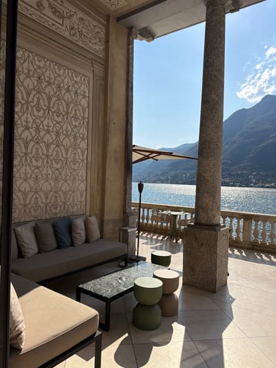 The image shows a shaded outdoor seating area overlooking a body of water and mountains. In the foreground, there is a long, upholstered sofa with several cushions, accompanied by a rectangular coffee table with a marble top and a dark metal frame. Next to the coffee table are three round stools of varying colors: green, beige, and brown. The seating area is on a tiled patio, protected by a stone column and an overhead structure.
Beyond the patio, a white balustrade lines the edge of a terrace. On this terrace, there are small tables and chairs, and a large umbrella provides shade. The water is sparkling under the sunlight, and mountains rise in the background, dotted with buildings. The sky is clear and blue, with a few white clouds. The overall scene suggests a luxurious and serene setting, possibly a hotel or a private residence in Pognana Lario, Italy, given the characteristic lake and mountain landscape.
