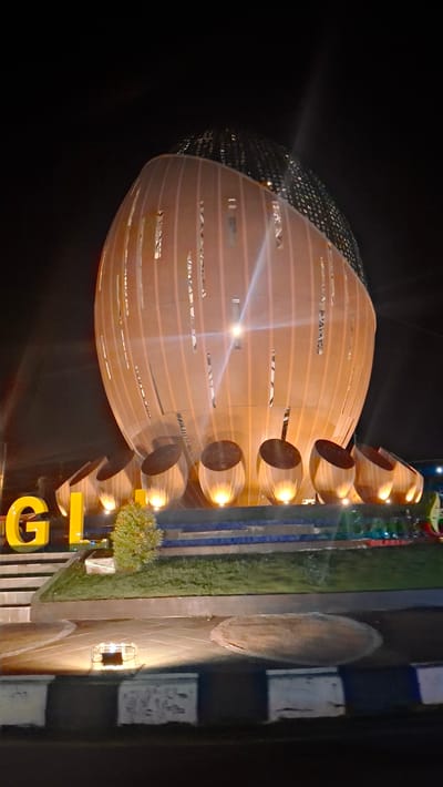 The image shows a nighttime view of the Tugu Bambu Runcing monument in Lampeudeu Baroh, Indonesia. The monument, shaped like a large, stylized bamboo shoot, is illuminated by spotlights, casting a warm orange glow. Its cylindrical body features vertical slits, and a patterned dome crowns its apex. Surrounding the base are several smaller, rounded structures, also lit from within. In the foreground, large yellow letters spelling "GL" are visible, along with a small, green bush. The monument is situated on a roundabout with paved paths and grassy areas. The scene is captured at night, with the dark sky providing a backdrop.