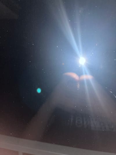 This is a photograph of a dark surface, likely a screen, with dust particles visible. There is a bright light source in the upper right quadrant of the image, creating lens flares and streaks of light. In the lower half of the image, a blurred reflection of what appears to be two hands or an object resembling hands is visible. A faint turquoise-colored circular anomaly is present in the left half of the image, possibly a reflection or a light artifact. The bottom edge of the frame shows a lighter area, suggesting the presence of a surface below the main subject. There is no discernible information about the location in Bašť, Czechia, as the image lacks any identifying landmarks or contextual elements.