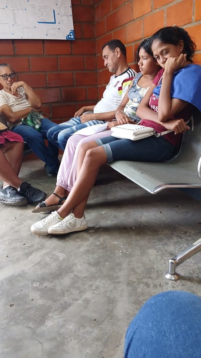 A group of people is seated on and around a metal chair in what appears to be a waiting area. Two young women are seated on the chair, with another woman standing behind them, her arms around their shoulders. A man wearing a white shirt with the Venezuelan flag on the shoulder is sitting next to the chair. An older man with glasses is seated on the floor to the left, his arms crossed. The floor is concrete and has a cracked surface. A brick wall is visible in the background on the left side of the frame. A poster with a schematic diagram is hanging on the brick wall. The location is assumed to be in Barcelona, Venezuela.