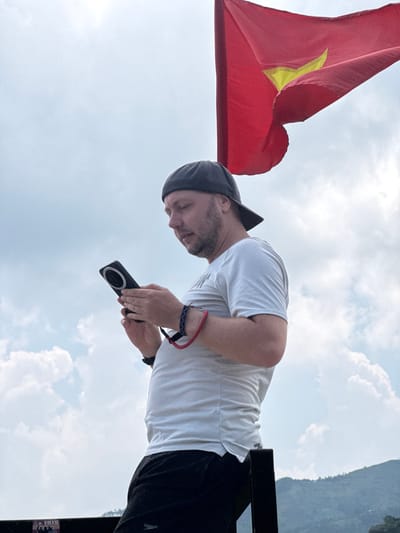 A person stands outdoors in Lũng Phìn Commune, Vietnam, holding a black electronic device with a circular detail. They wear a white t-shirt, black bottoms, a dark cap, and wrist accessories. A red flag with a yellow star is prominent to the left, and a cloudy sky fills much of the background. Part of a mountain or hill is visible in the upper right.