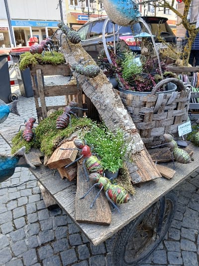 A wheeled wooden display unit occupies a cobblestone street in Homburg, Germany, positioned in front of commercial buildings. The unit features an elaborate arrangement of natural elements and artificial creatures. Seven large, stylized ant sculptures, varying in color from red-brown to green-and-red striped, are distributed across the display. They are placed on sections of bark-covered logs, clumps of green moss, and various pieces of cut lumber. A prominent, textured log spans diagonally across the display surface.

Potted plants, including specimens with small white flowers and others with dark red foliage, are interspersed within the arrangement. Some plants are contained within a large, woven wicker basket integrated into the display. A blue ceramic object, shaped somewhat like a stylized bird or fish, is positioned above the display, functioning as a fountain with water visibly flowing from it into the lower sections. Adjacent to this, a small bird feeder with a blue roof is also present. A small white sign bearing the German word "AMEISE" (ant) is visible. On the lower left, a blue fish-shaped sculpture is partially visible.

The surrounding environment consists of dark grey cobblestone paving. In the immediate background, a black SUV is parked, partially obscuring storefronts. One shop sign is partially visible, displaying the German word "Steuer" (tax). Grey refuse bins are present to the left of the display. A person's lower legs, clad in blue trousers, are partially visible behind the parked SUV.