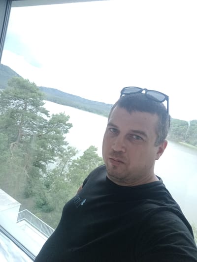 A man, wearing a black t-shirt with a blue logo on the left sleeve, stands indoors, looking directly at the camera. He has short, dark hair and is wearing sunglasses perched on top of his head. Behind him, through a large window, a lake and trees are visible, suggesting a scenic, possibly lakeside location. The man appears to be in Doksy, Czechia. The lighting is bright, indicating it is daytime.