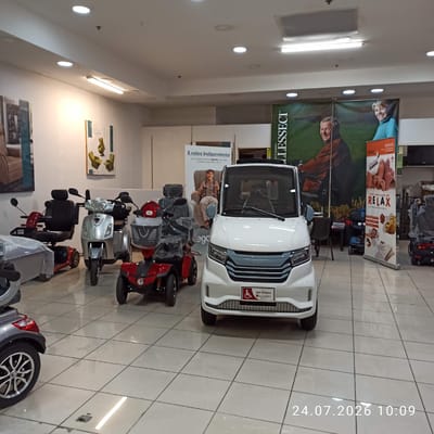 The image shows the interior of a retail showroom, likely a mobility scooter and electric vehicle dealership, located in Modugno, Italy. The showroom features a polished tiled floor and fluorescent lighting. Several electric mobility scooters and a white electric vehicle are displayed. A large banner with images of people and text in Italian is visible in the background, promoting a company called "ELLSSECI". A smaller banner with similar branding and product images is also present. The date stamp on the image indicates July 24, 2026, at 10:09 AM.
