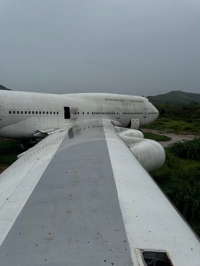 An abandoned Boeing 747 airplane rests in a grassy, overgrown field under a cloudy sky in Pak Nam Pran, Thailand. The aircraft, a dull white with visible dirt and grime, is partially obscured by another wing in the foreground, which is also weathered and discolored. The passenger cabin of the visible 747 shows rows of small windows, some of which are dark. One door is ajar, revealing an open hatch. The surrounding environment consists of dense green foliage and rolling hills in the distance, suggesting a rural or remote location. There are no people visible in the image.