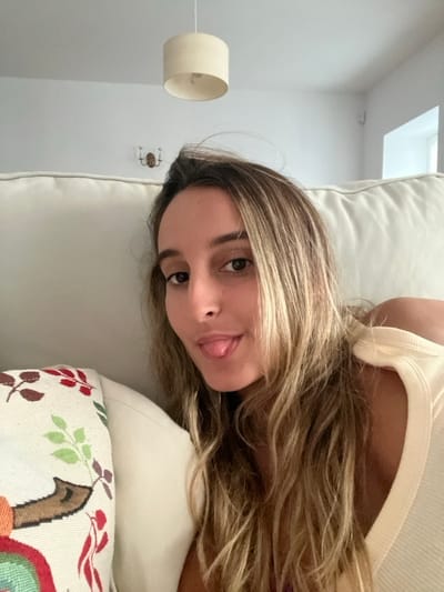An indoor photograph captures a young adult female from the chest up, leaning against a light-colored sofa. The female has long, wavy, light brown hair with blonde highlights, dark eyes, and is looking directly at the camera with her tongue slightly extended. She wears a light-colored, possibly cream, ribbed top. To her immediate left, a decorative throw pillow with a pattern of green, red, and brown leaves is visible on the sofa. The background consists of white walls and a white ceiling. A cream-colored cylindrical ceiling light fixture is suspended from the ceiling. A two-arm, brass-toned wall sconce is affixed to the wall in the upper left background. A portion of a white window frame is visible in the upper right background. The specific location within the city of Oliveira de Frades, Portugal, cannot be determined from the visual information presented.