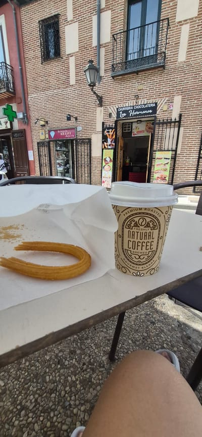 In Navalcarnero, Spain, a close-up shot captures a table with a churro and a disposable coffee cup. The churro, a U-shaped fried dough pastry, rests on white paper. Next to it, a beige coffee cup displays the text "FRESHLY NATURAL COFFEE BREWED GOURMET BLENDS." In the background, the facade of a brick building hosts a sign that reads "CHURRERIA CHOCOLATERIA Las Hermanas." The building features windows with wrought-iron bars and a lamppost. To the left, another storefront displays a green cross symbol, indicating a pharmacy. The ground is covered in small, light-colored pebbles. A person's leg and foot wearing a sandal are visible in the lower right corner of the frame.