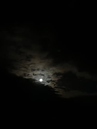 A night scene shows the moon partially obscured by clouds. The moon is a bright, circular object near the center of the frame. The clouds appear dark and diffuse, surrounding and partially covering the moon. The background is completely black. This is likely a nighttime photograph taken in Gorna Kremena, Bulgaria.