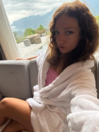 A woman with curly brown hair is sitting on a gray upholstered chair or sofa, wearing a pink top and a white terrycloth bathrobe. The scene overlooks an exterior space with stone tiles, a small planting box with greenery, and a mountainous landscape under a cloudy sky, characteristic of Cat Cat Village, Vietnam. The woman is looking toward the camera with a relaxed expression.