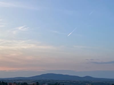 The image captures a serene twilight sky over a rural landscape, likely in Arandjelovac Municipality, Serbia. The upper portion of the sky is a clear, pale blue, transitioning to softer hues of pink and lavender near the horizon, indicating the sun has recently set. wispy clouds are scattered across the sky, and a faint contrail from an aircraft stretches diagonally from the upper right towards the center. Below the sky, rolling hills are silhouetted against the fading light. In the foreground, hints of a village or settlement are visible, with dark rooftops and trees dotting the landscape. The overall atmosphere is peaceful and expansive, with a focus on the natural beauty of the sky and land. There are no people or animals visible in the image.