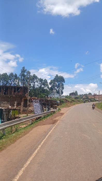 The image depicts a semi-urban roadside scene in Boochi Tendere ward, Kenya, under a clear blue sky with scattered white cumulus clouds. A two-lane asphalt road, light grey-brown with a faded white edge line on the left, curves gently to the right and recedes into the distance. Reddish-brown dirt shoulders flank both sides of the road.

On the left, a silver-grey metal guardrail extends from the foreground into the mid-ground, bordering a grassy and earthy bank. Beyond this, a partially constructed or demolished two-story brick building with an exposed wooden roof frame and open window apertures is visible. Adjacent to this structure are smaller, makeshift stalls or shelters constructed from corrugated metal and wood. Behind these, a dense cluster of tall, slender trees with light green foliage obscures a white-walled building in the background. Scattered debris and building materials are present on the ground.

On the right side of the road, the reddish-brown dirt shoulder continues, bordered by an elevated earth bank with sparse vegetation. In the mid-ground, a single individual is operating a dark-colored motorcycle, traveling away from the camera in the right lane. The individual wears a light-colored upper garment and dark lower garments. Further down the road, on rising terrain, several multi-story buildings of varying construction are visible, indicating a developed area. Multiple utility poles with overhead power lines are positioned along this side of the road. One small dark object, possibly a bird, is visible in the upper right sky.