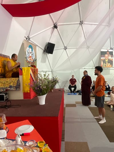 The image depicts an event, possibly a ceremony or performance, taking place indoors under a large white dome-like structure with visible support beams. The location is suggested to be in or around Mekića Kula, Montenegro.
Several individuals are visible. A man in an orange shirt and grey shorts stands with his head bowed, followed by a person in maroon monastic robes, and another individual in a maroon shirt sitting on a blue mat. Further in the background, more people are seated.
In the foreground, a monk in yellow robes is sitting on the floor with his hands in a prayer position, wearing a headset microphone. He is seated next to a red-and-brown-covered raised platform. On the platform is a vase of flowers, some religious objects covered in yellow and orange fabric, and a table with various objects including plates, cups, and other unidentified items. A speaker is attached to the white dome structure above the platform.
Other elements include decorative red fabric hanging from the dome structure and a poster of what appear to be religious figures. The floor is covered with carpet with a repeating pattern.