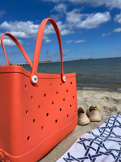 The image, which appears rotated 90 degrees clockwise, captures a sunny beach scene in Pionersky, Russia. A large, bright orange Bogg Bag dominates the foreground, extending from the bottom right to the mid-left. Its distinctive features include numerous circular perforations and white circular fasteners on the handles, each bearing a black lowercase 'b' logo.
To the right of the bag, a white towel with a navy blue geometric pattern is laid out on the sand. Next to the towel are a pair of light beige, perforated sandals, possibly Crocs, with one shoe containing a small golden-colored object.
The central part of the image shows calm, greyish-blue water, likely part of the Baltic Sea, stretching towards the horizon. The left side of the frame is filled with a bright blue sky, adorned with scattered white, fluffy clouds. In the far distance, faint silhouettes of several tall industrial cranes are visible above the waterline, suggesting a port or coastal construction in the vicinity of Pionersky. The scene depicts a bright, clear day, inviting relaxation by the sea.