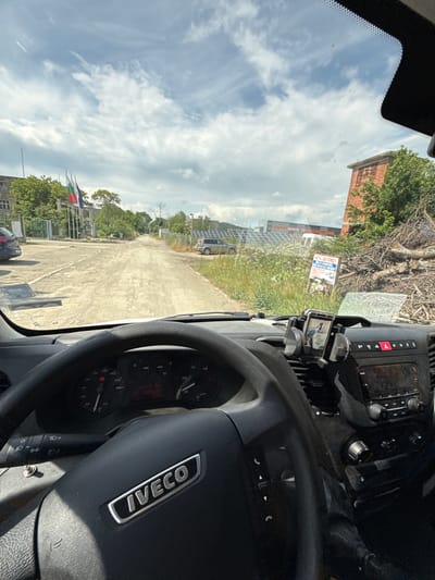 The image captures a driver's perspective from inside an Iveco commercial vehicle, looking out through the windshield on a bright, partly cloudy day. The original image is rotated 90 degrees, so viewing it upright shows the steering wheel on the right, indicating a left-hand drive vehicle.
The interior of the vehicle reveals a black steering wheel with a metallic "IVECO" logo. Behind it, the dashboard instruments, including a speedometer and tachometer, are partially visible. To the right of the steering wheel, the center console features an infotainment system with a screen and various knobs and buttons, including a prominent red hazard light button. A smartphone is mounted in a holder to the right of the console, displaying an image of what appears to be a bird or small animal.
Outside the vehicle, a concrete or dirt path extends into the distance. To the left of the path, a red-brick building is partially seen through trees, and further back, another industrial-looking building. A large array of solar panels is installed along this side. A white sign in Bulgarian is clearly visible, displaying "MUTA - ЖИВОТНОВЪДСТВО" (MUTA - LIVESTOCK FARMING), with additional text such as "ПРОДАЖБА" (SALE) and "МЛЯКО" (MILK).
On the right side of the path, several flagpoles display flags; one has vertical red, white, and green stripes, another is blue, and a third is red, all partially obscured by green foliage. A dark-colored car is parked in the foreground near the flags. The sky is bright with scattered white clouds, suggesting mid-day. The scene depicts a semi-rural or industrial area, consistent with the location context of Kalitinovo, Bulgaria, hinting at an agricultural or farming facility.