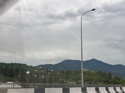 The image shows a view of a green, hilly landscape with scattered houses, likely in the city of Chakvi, Georgia. The sky is overcast with gray clouds, creating a muted light. In the foreground, there is a road barrier with black and white stripes. A tall, slender lamppost stands to the right of the frame, its curved arm holding a single light fixture. The hills are covered with dense green vegetation, including trees and bushes, and several houses of various sizes and architectural styles are visible nestled among them. In the background, a larger mountain range looms under the cloudy sky. The scene appears to be captured from a moving vehicle, given the slight motion blur. No people are visible in the image.
