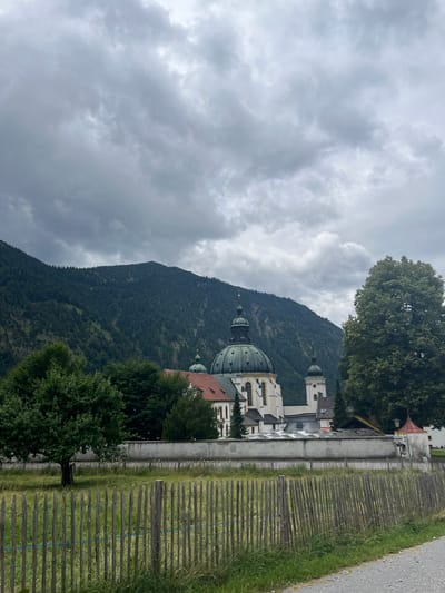 The image shows the Ettal Abbey in Ettal, Germany, under a cloudy sky. The large monastery complex, characterized by its Baroque architecture and a prominent green dome, is situated at the foot of a densely forested mountain. In the foreground, a rustic wooden fence runs across a grassy field. A solitary tree stands to the left of the frame, while a larger, mature tree is positioned to the right, partially obscuring the monastery's bell tower. The overall scene conveys a sense of tranquility and historical significance, with the imposing natural landscape of the Bavarian Alps serving as a backdrop to the architectural marvel.