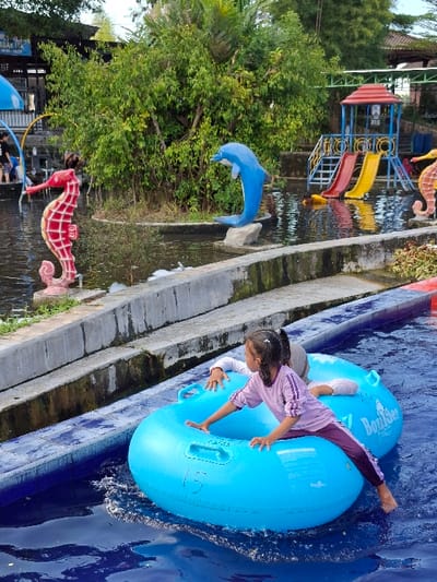 The image shows a scene at a water park in Kecamatan Tulung, Indonesia (latitude: -7.592, longitude: 110.65). Two young girls are seated in a blue inflatable raft, floating on a pool of water. The girls are wearing casual clothing. The raft is marked "Boogie". The girls are in the water, and the scene is set at an outdoor water park. The scene is bright and sunny. Behind the girls, there are several decorative objects, including a blue dolphin statue, a red and yellow seahorse statue, and a playground structure with a slide. There are lush green trees and shrubs in the background. A concrete wall surrounds the pool.
