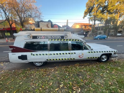 The image shows a parked, white, modified 1959 Cadillac Miller-Meteor ambulance, replicated to resemble the "Ecto-1" vehicle from the Ghostbusters franchise. The car is parked on a street lined with buildings and trees, and foliage on the ground suggests autumn. The vehicle has Ghostbusters branding on its sides and top, along with hazard stripes and a logo depicting a ghost. The text "ECTO1 ARGENTINA 11 6806,9768" and "WE BELIEVE YOU" call number on its side indicates it is located in Argentina. The setting appears to be a street in the city of Martínez, in the province of Buenos Aires, Argentina. There is also a blue car visible in the background.