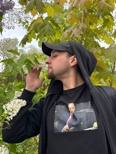 A man in a black hooded jacket and a black cap is standing outside, seemingly smelling or looking at white lilac flowers on a bush. The jacket has a print of Vladimir Putin on the front. The background includes green maple leaves and more lilac bushes. The location is unspecified.
