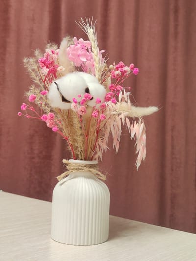 A vertical, medium shot captures a bouquet of dried flowers and cotton in a white ribbed vase. The vase is adorned with a twine tie around its neck. The bouquet features fluffy white cotton balls, delicate pink flowers, and wispy dried grass and seed pods in various shades of tan and pink. The background is a muted, textured reddish-brown curtain. The vase rests on a light-colored, wood-grain textured surface, which is a table. The scene appears to be indoors, possibly in Guryevsk, Russia, given the prompt's inclusion of that location. No people or actions are depicted.
