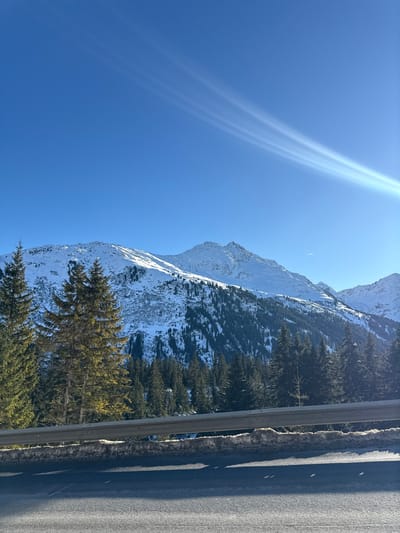 The image depicts a winter mountain landscape in St. Christoph, Austria, under a clear, bright blue sky. Snow-covered peaks and slopes dominate the mid-ground and background, with coniferous forests covering the lower mountain flanks. The foreground features a dark asphalt road surface, above which a metallic silver guardrail runs horizontally across the frame. Behind the guardrail, accumulated dirty snow and ice are visible, along with several tall, sun-illuminated coniferous trees. Multiple parallel light rays, indicative of sun glare, emanate from the upper right quadrant of the sky. No individuals, animals, or active human interactions are present.