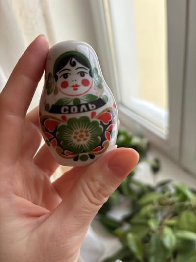 A light-skinned human hand holds a white ceramic salt shaker shaped like a traditional Russian matryoshka doll. The doll is adorned with a folk art design featuring a woman's face with black hair, green headscarf accents, red cheeks, and red lips. Below the face, a black band displays the Cyrillic word "СОЛЬ" (SOL), meaning "SALT," in white letters. The lower body of the shaker exhibits a large green floral pattern with red and green embellishments. The scene is illuminated by natural light entering through a window with a white frame in the background. Beyond the window, a light-yellow exterior is out of focus. Blurry green foliage is visible to the lower right. The specific location of this scene is in the city of Velikiye Luki, Russia.
