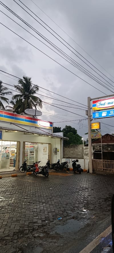 This image depicts an Indomaret convenience store and its immediate surroundings in Landangan, Indonesia, during overcast conditions at dusk or early evening. The scene is dominated by the store's white facade with its signature red, blue, and yellow horizontal striped canopy. The interior of the store is brightly lit, revealing aisles of products and one individual standing near the counter in a light-colored shirt and dark trousers. A second person's lower leg is partially visible further inside.

Nine motorcycles are parked on the wet, dark grey paver-stone ground in front of the store; three are closer to the foreground, and six are positioned nearer the entrance. The ground shows puddles and appears muddy in some areas. A tall vertical sign on the right displays the "Indomaret" logo at the top, followed by illuminated signs for "pulsa," "payment," and a "mandiri atm." A smaller, horizontal Indomaret sign is mounted above the store entrance.

Above the store, several palm trees are visible against an overcast, light grey sky. Numerous black utility lines crisscross the upper portion of the frame. Two white satellite dishes are affixed to the roof of the Indomaret building. To the right of the store, behind the parking area, a grey concrete block wall separates the property from dense green foliage. A street light stands illuminated to the right of the store, and a simple wooden fence precedes an obscured building further right.