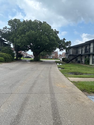 This is a photograph of an apartment complex and its surrounding grounds. The image depicts a paved road in the foreground, with a concrete walkway on the right. Beyond the walkway, a grassy area with several large puddles is visible. Two-story apartment buildings with balconies and outdoor staircases are situated on the right side of the image. A large, mature tree with dense green foliage stands prominently in the middle ground, casting shadows on the road. Other trees and landscaping are visible throughout the scene, along with a few parked vehicles in the distance. The sky above is partly cloudy, with patches of blue and gray. The overall environment suggests a residential setting within a city, likely Balch Springs, United States of America, possibly after a rainfall given the puddles.