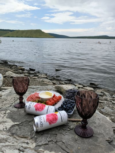 The image depicts an outdoor scene on the rocky shoreline of a large body of water in Horaivka, Ukraine, under a partly cloudy sky.
In the immediate foreground, a picnic setup is arranged on a large, flat, grey-brown rock. This includes two dark purple, textured glass goblets positioned on either side of the setup. Central to the arrangement is a white circular plate holding slices of cured sausage, a small white bowl containing a yellow creamy substance, several red cherry tomatoes, and green pickled gherkins. Adjacent to the plate are a clear plastic container filled with blueberries and a small bundle of dried purple flowers, resembling lavender, tied with natural twine. Two white aluminum cans, identified as "Spragg Kombucha" with "вишня" (cherry) and "малина" (raspberry) flavors, lie horizontally near the food.
The midground features the edge of the rocky shoreline, composed of various grey and brown stones, giving way to the calm, rippling surface of the water. Several indistinct figures are visible in the water in the mid-distance, appearing to be swimming or wading.
In the background, across the expanse of water, are rolling green hills under a light blue sky with scattered white cumulus clouds. The farthest hills appear darker due to distance.