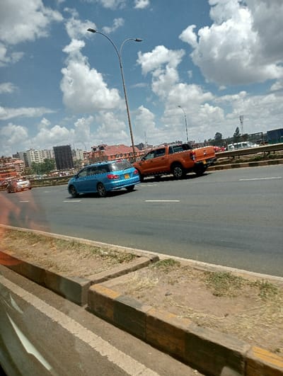 A blue hatchback and an orange pickup truck travel on a paved highway, separated by a white line. Lampposts line the side of the road. A concrete median with patches of grass sits between lanes. Buildings and a cloudy sky with patches of blue form the backdrop. The image appears to be taken from another vehicle.