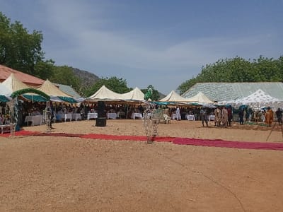 The image shows a large outdoor gathering in Zum, Nigeria, located at the coordinates 10.116 latitude, 13.046 longitude. The event is taking place on a sandy ground, with several white tents set up, likely for a wedding or other celebration. There are many people present, mostly dressed in traditional attire. The scene features a red carpet leading to the tents, and there are some trees and a low-lying building in the background. The event appears to be well-organized, with tables and chairs set up under the tents. A large speaker system is set up in front of the main tent. The atmosphere is festive and crowded. The event is likely taking place during the day, as the sky is clear and bright.