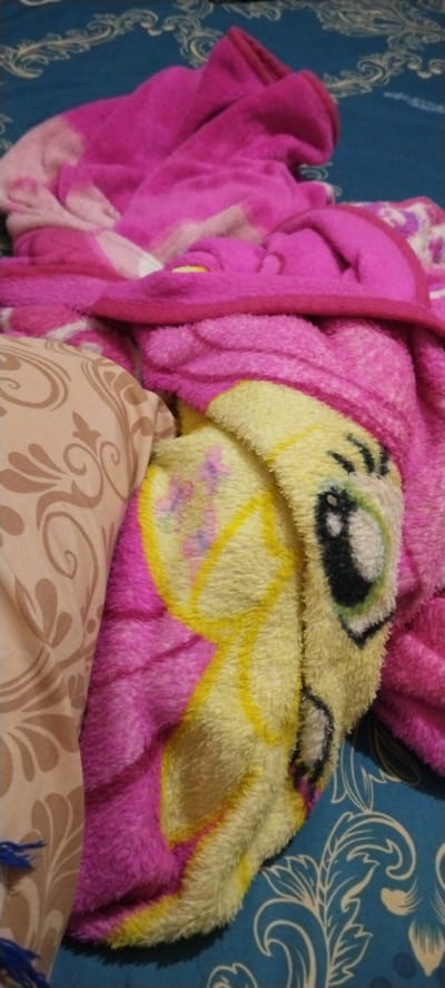 The image shows a close-up of a pile of soft, fluffy blankets, suggesting a cozy and domestic setting. The blankets are primarily pink and yellow, with one featuring a cartoonish character's face, possibly from a children's show. The character has large, round eyes with black pupils and a white outline, and its face is primarily yellow with some pink and purple accents. The texture of the blankets indicates they are likely made of fleece or a similar material, designed for warmth and comfort. One blanket has a light brown patterned fabric, possibly a pillowcase or another blanket, with a scrolling, floral motif in a darker brown. The background appears to be a blue patterned fabric, possibly a bedspread or sofa, with a stylized white floral design. The overall impression is one of comfort and perhaps a child's bedroom or a living space where these blankets are used for relaxation. The lighting is somewhat dim, suggesting an indoor setting, possibly at night or on a cloudy day. There is no visible text in the image. The scene appears to be set in Duman, Indonesia, based on the provided location context.