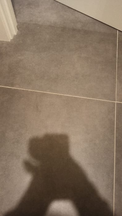 The image depicts a close-up of gray tiled flooring with white grout lines. A shadow of a person's hand and arm is cast on the tiles on the right side. The left side features white baseboards. The tiles have a matte finish with subtle variations in color.