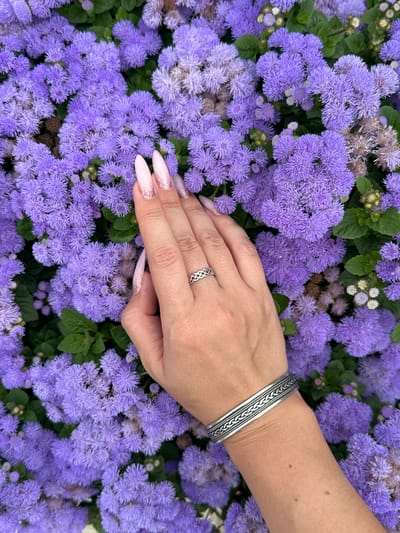 The image captures a close-up view of a human hand and forearm gently placed among a dense cluster of vibrant purple flowers, likely Ageratum (Flossflower). The flowers are soft, fluffy, and numerous, with various shades of purple and hints of green foliage peeking through. Some older, lighter-colored flower heads are also visible, indicating a natural growth cycle.

The hand, likely female, is fair-skinned. It features long, almond-shaped nails with a light pink base and sparkling, glittery tips. A silver-toned ring with an intricate, braided, possibly Celtic knot-like design adorns the ring finger. Further up the forearm, a wide, silver cuff bracelet is worn, featuring detailed braided and textured patterns along its surface. The forearm itself shows a few small, dark moles.

The activity depicted is the hand resting softly on or among the flowers, suggesting a moment of interaction, contemplation, or appreciation for nature. The lighting is bright and even, indicating a daytime setting. The scene conveys a sense of natural beauty and tranquility, set in what appears to be a cultivated garden or flower bed. The location context for this image is Buk-myeon, South Korea. There is no visible text in the image.