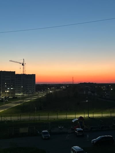 The image depicts a dusk or sunset scene in Klintsy, Russia. The sky transitions from blue to orange. In the foreground, there is a building under construction with a crane visible. Adjacent to the building is a road with streetlights. Further away, there is a parking lot with cars, and a playground. The area appears residential.
