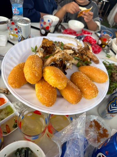 The image captures a meal being shared by several people at an outdoor dining setting, likely in Bình An, Vietnam, given the brand name of one of the beverages.

Center stage is a white plate featuring an assortment of food. On one side sits the remains of what appears to be fried fish or chicken, with bones and scraps evident. The other half of the plate is filled with golden-brown, oblong-shaped fried dough.

The table surrounding the plate is cluttered with various items. There are glasses with colorful polka dots, a can of beer, a water bottle, and small dishes containing sauces and condiments. Some people are seated around the table, one seen holding a piece of fried food and another a bowl.