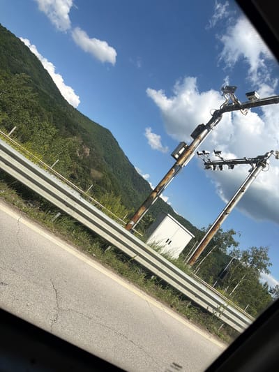 The image depicts a roadside scene in Vrachesh, Bulgaria. Two metal poles with traffic signals are prominently featured, positioned over a highway. The poles appear weathered and slightly rusted, suggesting they have been in place for some time. Attached to the poles are multiple traffic lights and what appear to be sensors or cameras, indicating an intelligent transportation system.
To the right of the poles, a white, rectangular electrical cabinet is visible, likely housing control equipment for the traffic signals. The highway itself runs diagonally across the frame, with a metal guardrail separating it from the grassy embankment. The embankment is covered in green vegetation and some wildflowers.
In the background, a densely forested mountain slope rises steeply, its greenery contrasting with the bright blue sky and scattered white clouds. The lighting suggests it is daytime, likely late morning or early afternoon, with clear and sunny weather. The perspective is from within a vehicle, looking outwards through a window, which is indicated by the dark framing on the edges of the image.
No people are visible in the image. The scene is quiet and appears to be a typical view from a vehicle traveling along a mountainous highway. There is no visible text in the image.