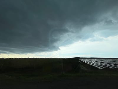 This media file depicts a rural scene in Davidovo, Bulgaria. The image's primary subject is a dramatic sky filled with dark, ominous storm clouds on the left, contrasting with lighter, broken clouds and a clear sky towards the right. Below the sky, a dark, silhouetted landscape stretches across the frame. In the mid-ground, a field with rows covered in plastic sheeting is visible on the right, suggesting agricultural activity. A small, dark structure, possibly a shed or shelter, stands in front of these covered rows. The left side of the mid-ground shows a darker, less distinct field with a line of trees or vegetation forming the horizon. The foreground is a dark, uncultivated area, likely the ground from which the photo was taken.
The weather appears to be stormy or just before a storm, with the dark clouds creating a sense of anticipation or unease. The time of day is difficult to pinpoint precisely due to the overcast conditions, but the light suggests it is likely late afternoon or early evening, when the sun is lower in the sky. The overall mood is somber and dramatic due to the intense cloud formations. There is no visible text in the image.
