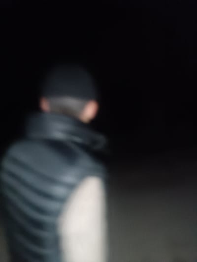 A severely out-of-focus image taken in Shamsikul, Uzbekistan, depicts the upper back of an individual in an extremely low-light environment, likely nighttime. The subject wears a dark cap or beanie and a dark, possibly padded vest over a lighter-colored garment, visible on the right arm/shoulder area. The individual faces away from the camera, looking towards the right of the frame. The background is featureless and uniformly dark, providing no additional environmental context. No specific actions or interactions are discernible.