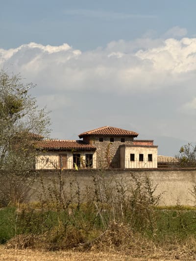 A stone building with a red-tiled roof stands against a sky filled with fluffy clouds. The building has windows and a tower-like structure with barred windows. To the right of the building, vegetation and a tall wall are visible. In the foreground, branches of a tree partially obscure the view of the structure. The overall scene suggests a rural or historical setting.