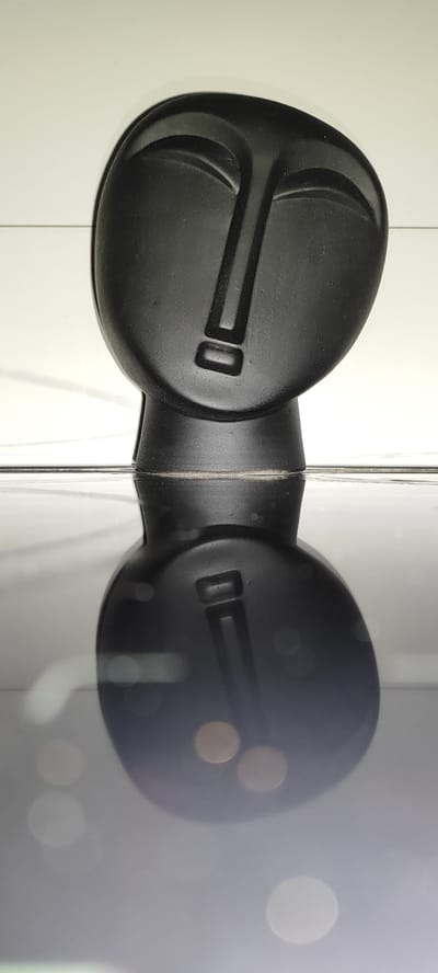 A black abstract sculpture of a face is positioned on a reflective surface, creating a mirrored image below it. The sculpture is dark, with carved features representing eyes, a nose, and a mouth. The background is a plain white surface, possibly a wall or shelf. The reflective surface below is dark and smooth, showing blurred circles of light, suggesting ambient lighting. The image is captured from a slightly elevated angle, looking down at the sculpture and its reflection. The location in Snezhinsk, Russia is not discernible from the visual information provided in the image.