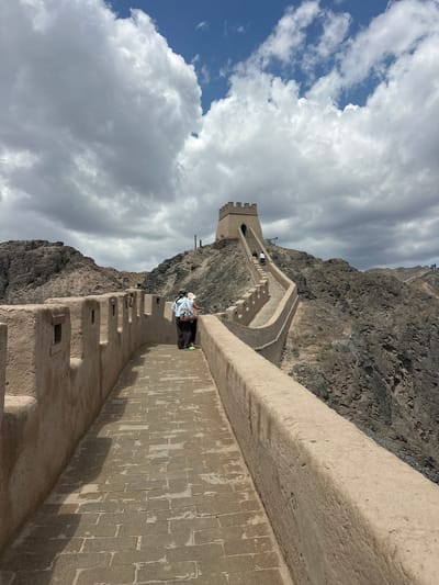 The image captures a section of the Great Wall of China near Jiayuguan, China. In the foreground, the broad, brick-paved walkway of the wall stretches into the distance. The wall itself is constructed of a sandy-colored material, with regularly spaced square openings in its parapet. Two individuals are standing on the walkway, facing away from the camera, with one person leaning on the wall. They appear to be tourists.
Further along the wall, the structure ascends a steep, rocky incline, culminating in a watchtower with a crenelated roof. Other individuals can be seen walking up the steep path towards the tower. The surrounding environment is arid and mountainous, with rugged, barren slopes on either side of the wall. The sky above is a mix of blue with large, white, fluffy clouds. The overall scene depicts a well-preserved section of the Great Wall under a dynamic sky, highlighting its architectural grandeur against a dramatic natural landscape.