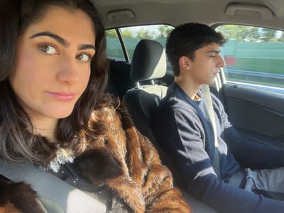 This image depicts two individuals inside a moving vehicle in Caminha, Portugal.
In the foreground, a female with long, dark, wavy hair and brown eyes looks towards the camera, slightly off-center to her right. She wears subtle makeup, including eyeshadow and lip gloss. She is dressed in a dark top featuring a white lace collar and a prominent brown, shaggy faux fur coat, with a light-colored seatbelt secured across her chest. Strong sunlight illuminates her right side, creating highlights on her hair and face.
Behind and to the right of the female, a male individual is seated, viewed in profile. He has short, dark hair and is looking forward, presumably at the road ahead. He wears a dark blue long-sleeved sweater and light grey trousers, with a seatbelt secured across his chest. The car interior features dark upholstery, and through the passenger window, a blurred green sound barrier or roadside foliage and clear sky are visible, indicating movement on a highway or open road. The sunlight casts distinct shadows within the vehicle, consistent with a sunny day.