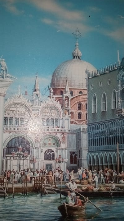 This is a painting depicting a bustling scene in Venice, Italy. The foreground features a canal with several gondolas. One gondola in the lower right has three people in it, one rowing and two passengers. Another gondola, larger and to the left, carries at least two people, one rowing. In the middle ground, a crowded boardwalk runs along the canal, lined with people engaged in various activities, some appearing to be merchants selling goods from baskets piled with produce. The background is dominated by grand Venetian architecture, including a large, ornate building with intricate stonework, arches, and statues, likely St. Mark's Basilica. To the right, a darker, more austere building with arched windows and a decorative facade stands. A large dome with a cross on top rises behind the basilica, suggesting it is part of a larger religious complex. The sky is a clear, bright blue with a few wispy clouds, and a couple of birds are visible. The overall impression is a lively, sunny day in Venice, with a vibrant atmosphere of commerce and daily life. The style of dress of the people suggests a historical setting, possibly the 18th or 19th century. There is no visible text in the image.