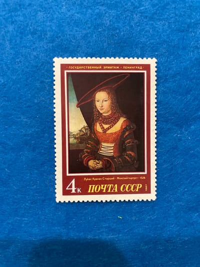 The image is a close-up, slightly angled photograph of a postage stamp. The stamp features a portrait of a woman in historical clothing, painted in a Renaissance style. The woman has fair skin, long, curly brown hair, and is wearing a red hat and a multi-layered, ornate dress with red and dark fabric. She is looking slightly to her left, with her hands clasped in her lap. Behind her, a landscape scene with buildings and a body of water is visible.
The stamp has a white border with serrated edges. Along the top of the stamp, in Cyrillic script, are the words "ГОСУДАРСТВЕННЫЙ ЭРМИТАЖ · ЛЕНИНГРАД ·" which translates to "STATE HERMITAGE · LENINGRAD ·". Below the portrait, there is a denomination of "4k" and further Cyrillic text: "ЛУКАС КРАНАХ СТАРШИЙ · ЖЕНСКИЙ ПОРТРЕТ · 1526", meaning "LUCAS CRANACH THE ELDER · WOMAN'S PORTRAIT · 1526". At the bottom right of the stamp, the text "ПОЧТА СССР" (POST OF USSR) is present, along with the year "1987".
The stamp is placed on a textured blue surface, likely fabric, which appears to be a felt-like material. There are no discernible elements in the image that indicate the location of Гамово, Russia. The stamp itself is a representation of artwork housed in the Hermitage Museum in Leningrad (now Saint Petersburg), Russia.
