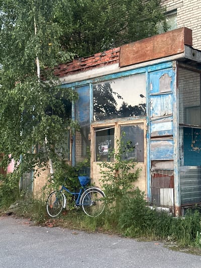 In the city of Kovalyovo, Russia, an abandoned structure stands weathered and overgrown. A blue bicycle rests against the crumbling facade, its tires resting on a patch of green grass and scattered debris. The building's blue-painted walls are peeling, revealing layers of faded paint and weathered wood underneath. Large, grimy windows reflect the surrounding trees, their glass cracked and broken in places. Overgrown bushes and trees encroach on the structure, their branches reaching towards the decaying roofline, which is capped with rusted metal. A brick chimney is visible on the left side of the building, partially obscured by foliage. The scene is set against a backdrop of a brick building, also showing signs of disrepair. The ground in front of the structure is a grey asphalt road, suggesting an urban or semi-urban environment. The overall impression is one of decay and neglect, with nature slowly reclaiming the man-made structure.