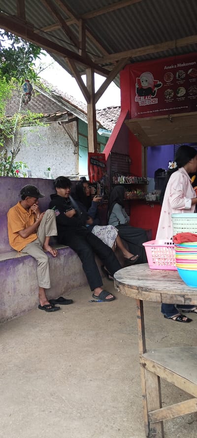 The scene takes place in a small, open-air restaurant or food stall located in Cimaung, Indonesia, at coordinates -7.074 latitude and 107.547 longitude.  Four people are seated at a wooden table, facing the camera. The person on the left is wearing a yellow shirt and beige pants. The person in the center is wearing a black jacket and black pants. The person to their right is wearing a floral dress and black pants. The person on the far right is wearing a pink dress and a white headscarf.  In the background, there is a counter with a sign reading "Aneka Makanan" and a variety of food items. There are also several brightly colored plastic containers stacked on top of each other, along with a pink wicker basket. The wooden frame of the restaurant structure can be seen above the table. The floor is made of concrete.  A green plant is visible outside the restaurant, with a small red flower on the ground nearby.  The overall mood of the scene is casual and relaxed. The people seem to be enjoying their meal and their surroundings.