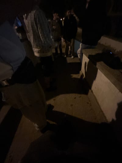 The image depicts a low-light, outdoor scene at night in Santa Maria da Feira, Portugal. At least five individuals are present on a paved surface, which appears to be a street or plaza. A light-colored concrete barrier or bench runs along the right side of the frame.

In the foreground left, a person is partially visible, wearing a light-colored top and dark trousers. In the mid-ground left, an individual wears a light-colored, possibly patterned, jacket or sweatshirt. Further back and to the center, two individuals stand; one is dressed in a dark jacket and lighter trousers, appearing to look at a small object in their hands, while the other also wears a dark jacket and light trousers. On the right, a person is seated on the concrete barrier, wearing a dark top and light pants, facing away from the viewer.

On the concrete barrier, a dark object with two distinct blue lights is present. The overall lighting is dim, indicating ambient night lighting, with some distant light sources visible in the background. There are no discernible specific interactions between the individuals beyond their proximity in a casual gathering.